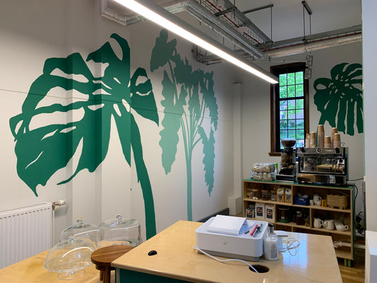 A café with a large dark green cheese plant leaf and mint green tropical leaves painted on white walls. In the foreground is a till and the background shows a coffee machine and storage.
