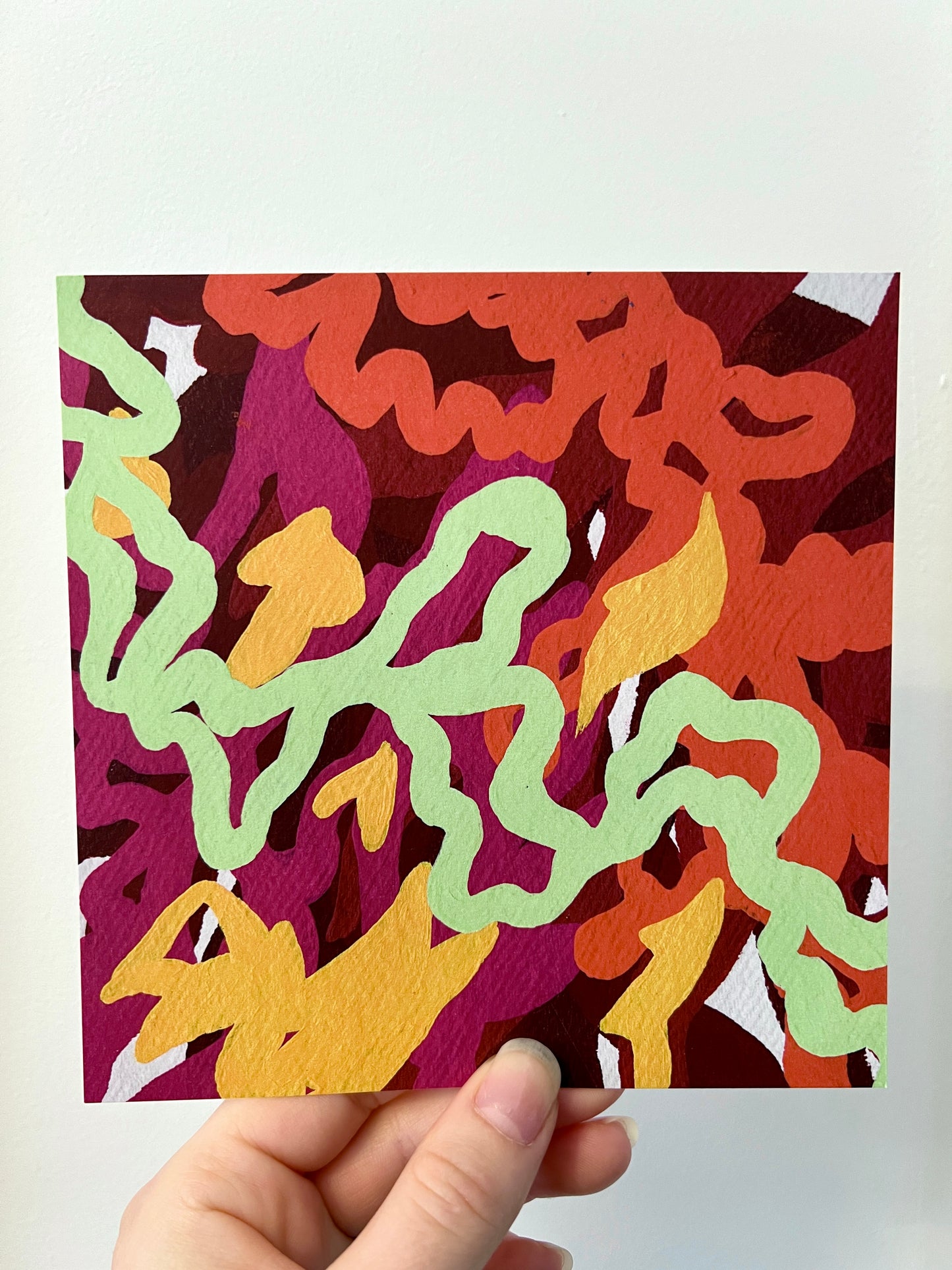 A hand holding a colourful print of a painting against a white wall. The print is layers of orange, red, pink and yellow abstract shapes with a light green swirling line across the image.