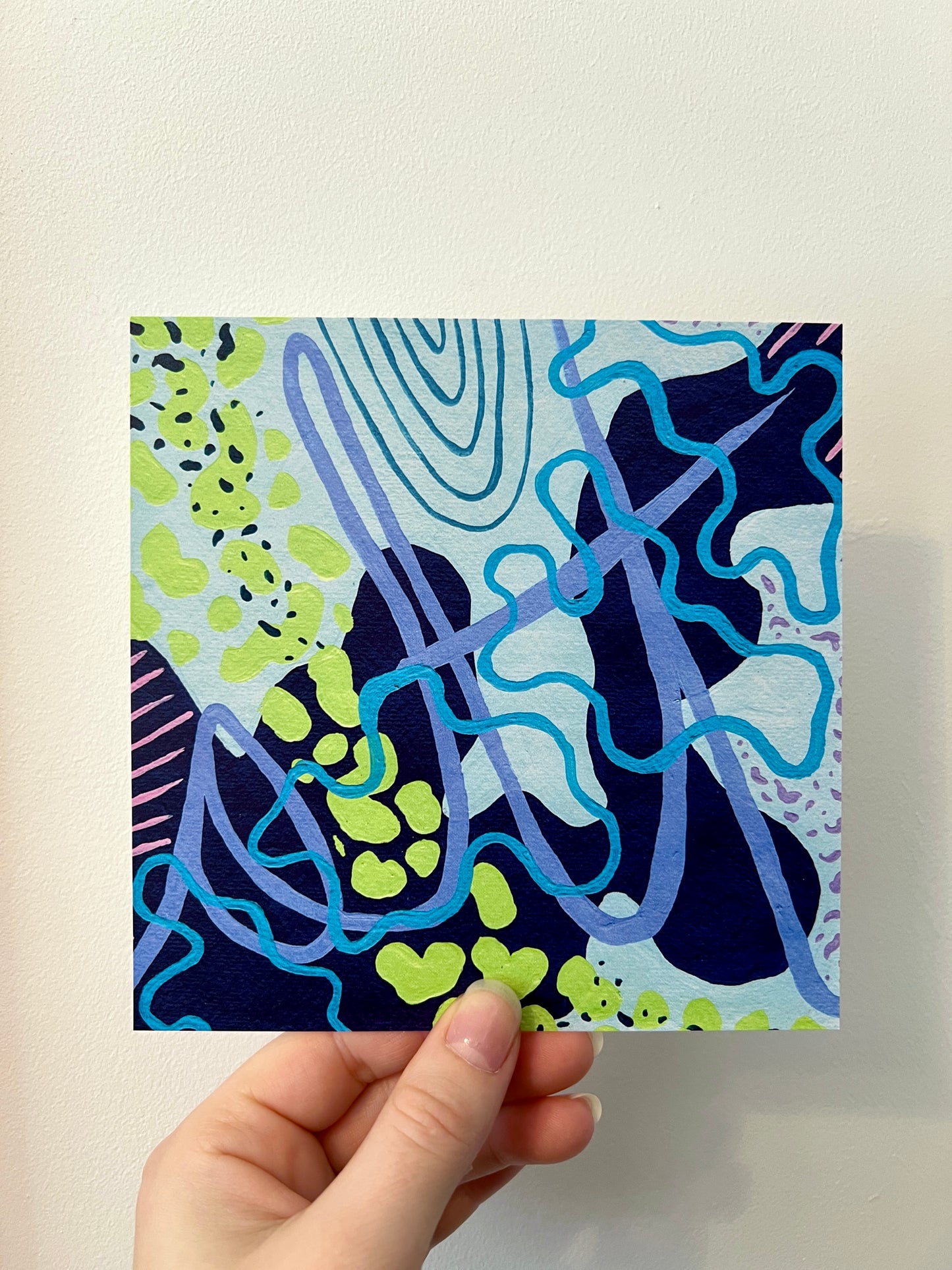 A hand holding a print containing abstract shapes of varying shades of blue with green shapes sweeping across against a white wall.