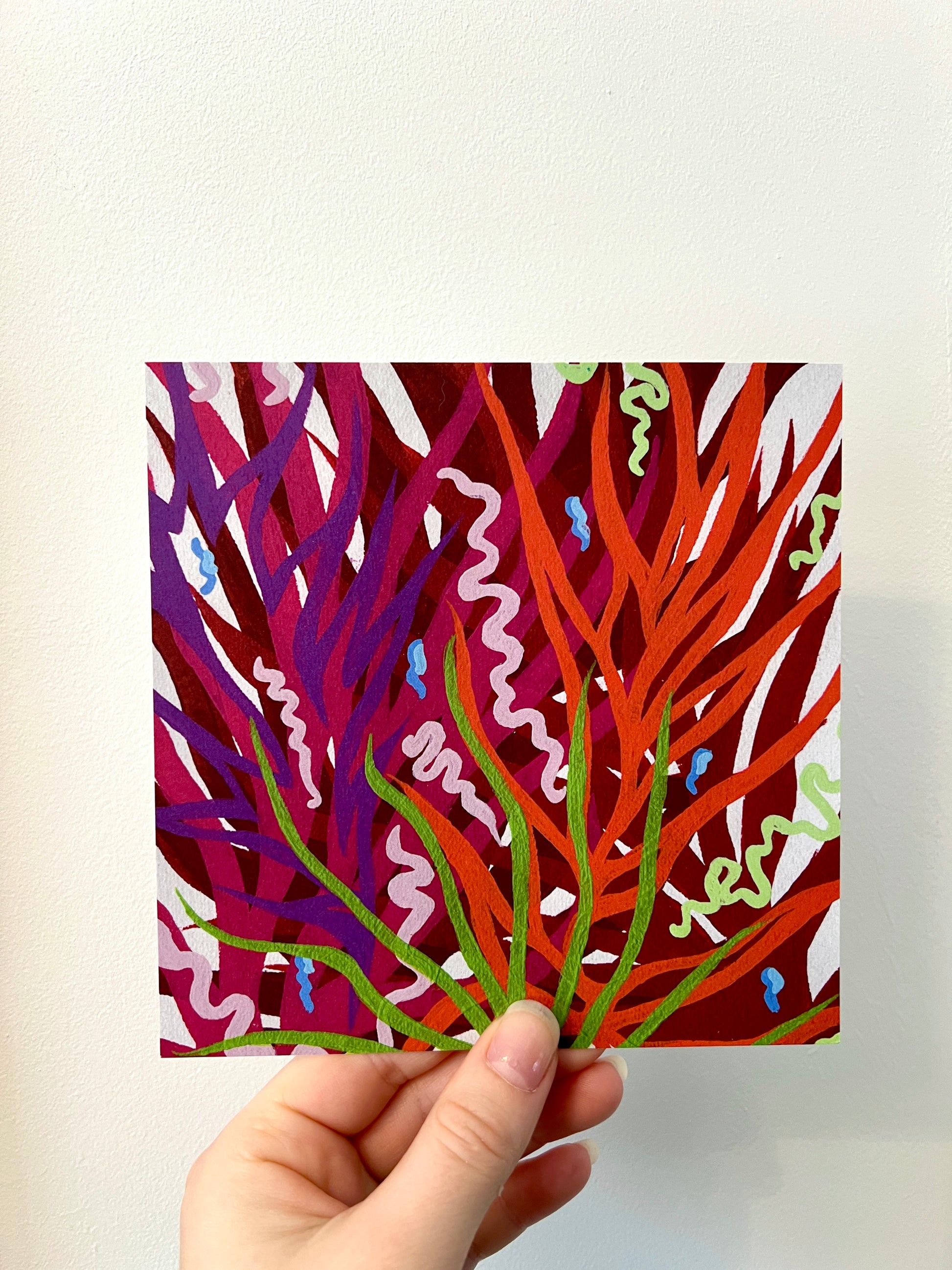 A hand holding a vibrant abstract print in front of a white wall. The print has plant like shapes in bold colours that look like they are growing up the design.