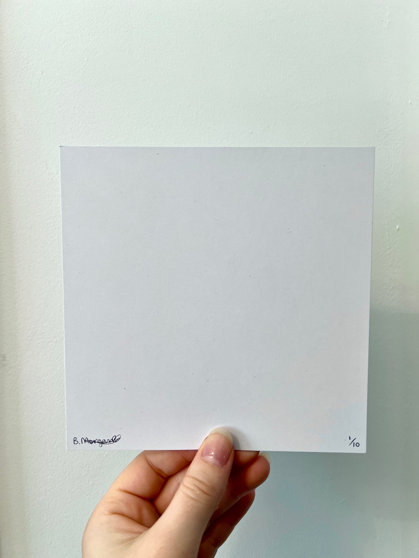 A hand holding the back of a print against a white wall. The print is white and square with a signature in the bottom left corner and a number in the bottom right corner.