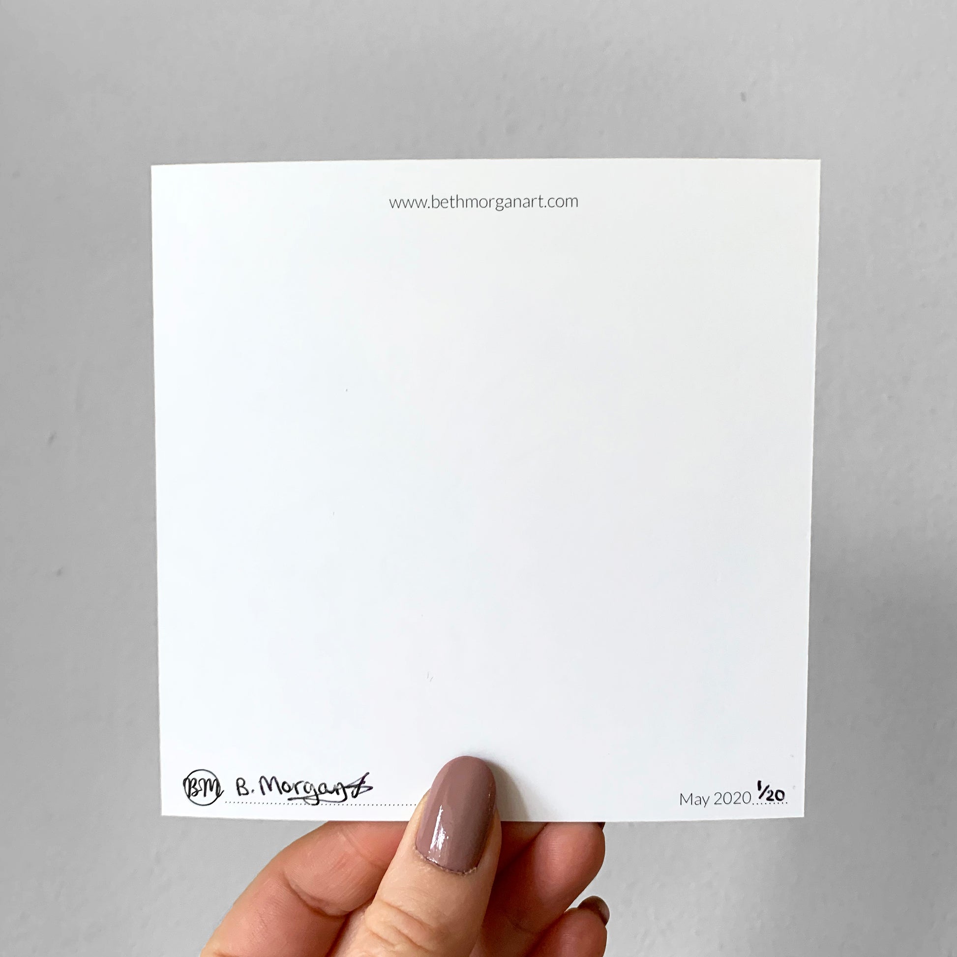 A hand holding a white square print which has been signed and numbered by Beth Morgan. It also includes a website address and a logo.