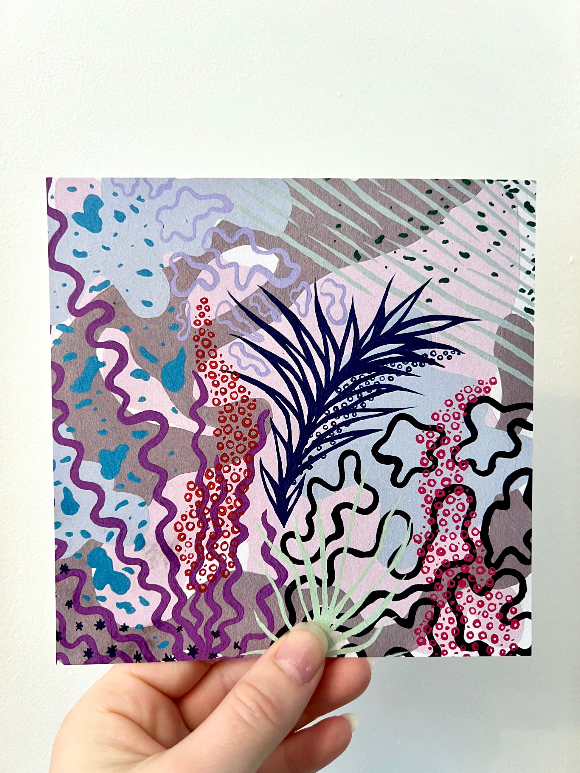 A hand holding a square print against a white wall. The print has shades of purple and pink with red and bright pink circles forming shapes like fox glove flowers. There are swirling lines and outlined shapes across the image.