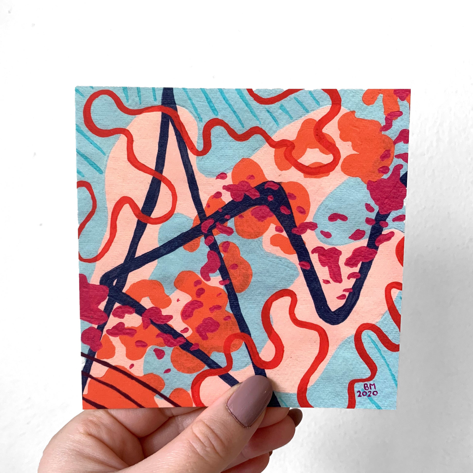 A hand holding a print of an abstract painting against a white background. The painting is made up of shades of blue and orange, and has pink shapes cutting across the diagonal along with a navy sharp line and red swirling lines. 