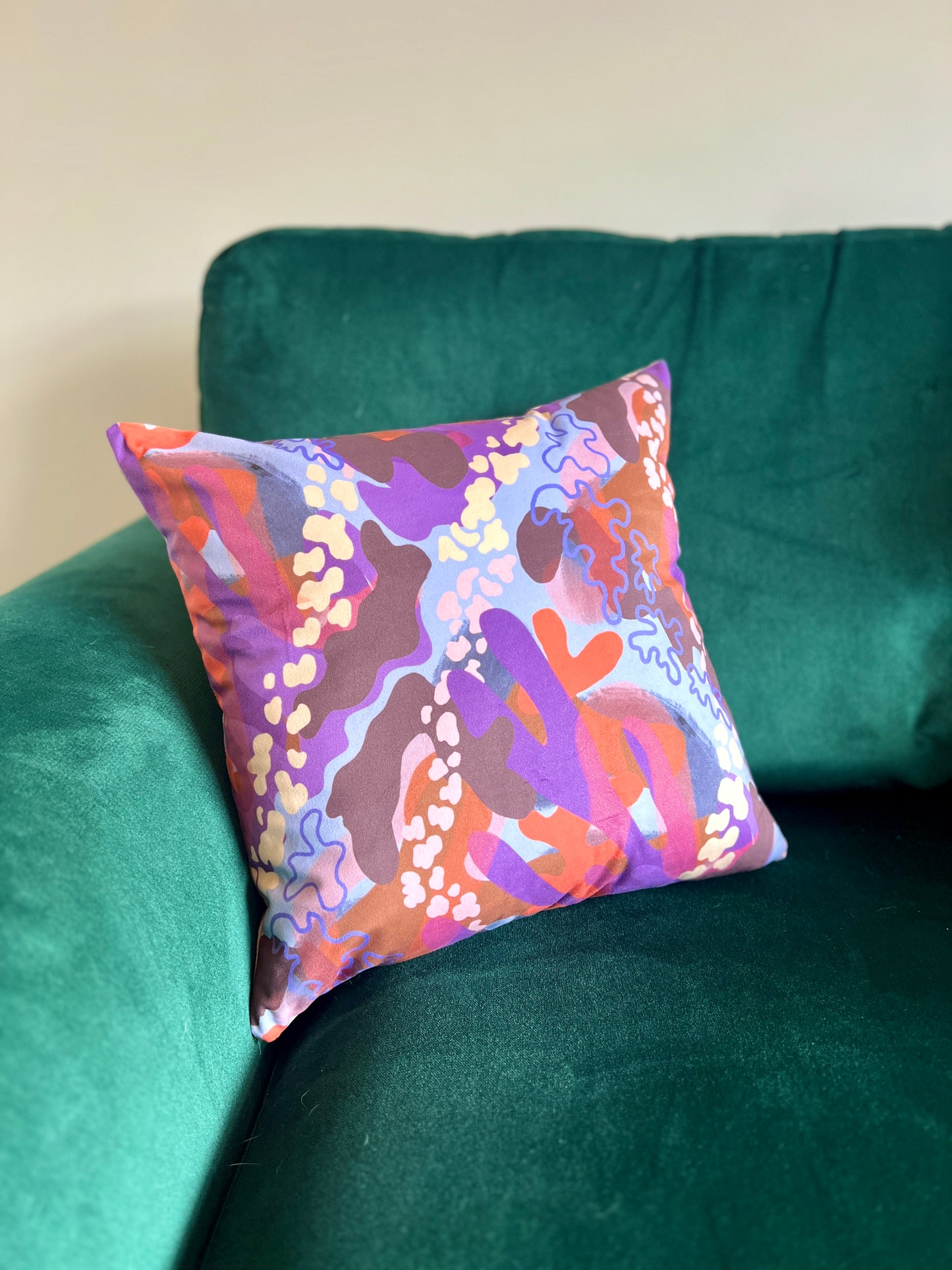 An abstract statement cushion placed on a deep green sofa. The cushion is made up of shapes of purple, orange and pink.