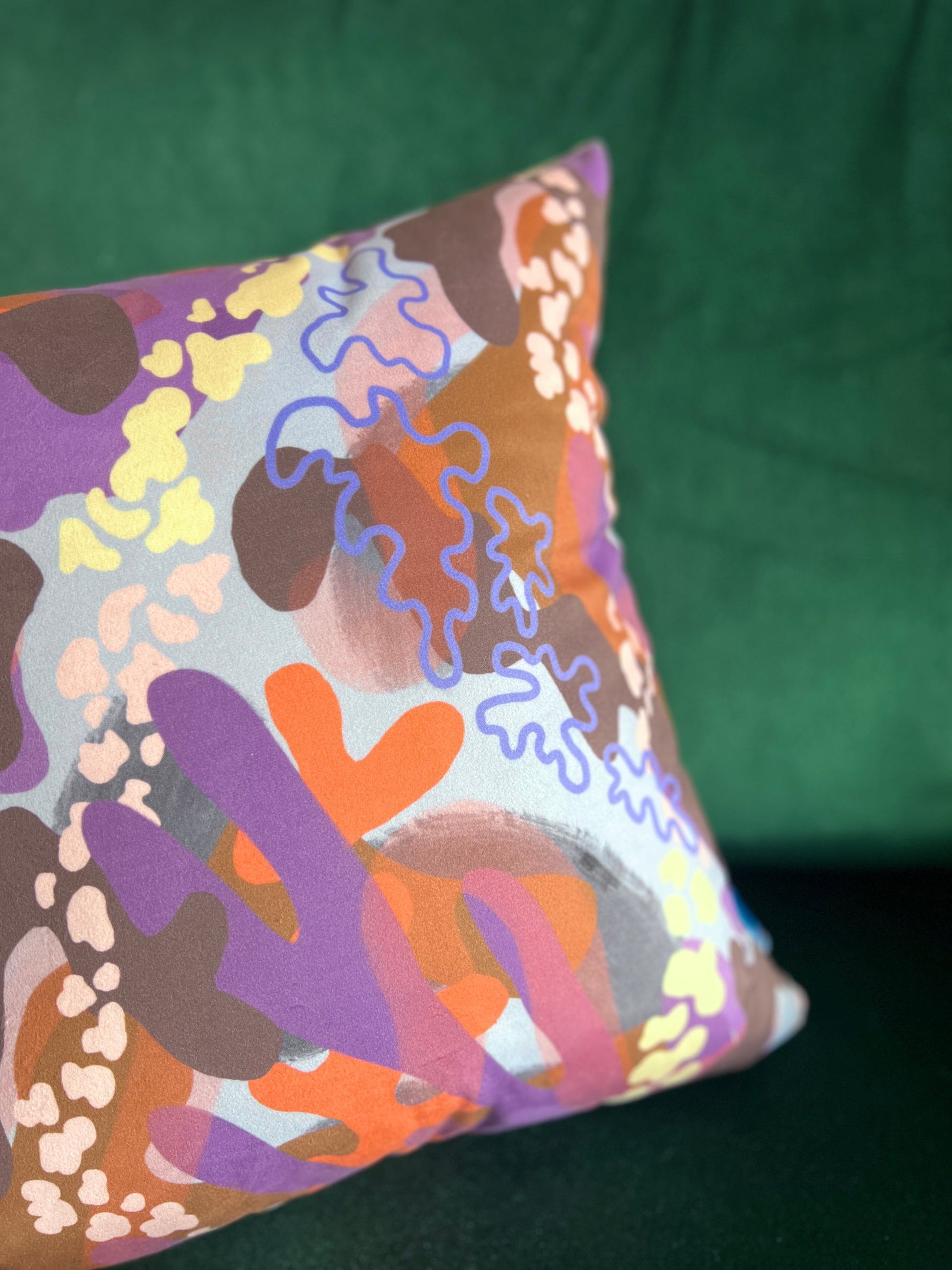 A close up of an abstract cushion on a green background. The cushion features abstract shapes and patterns in shades of purple, orange and pink.