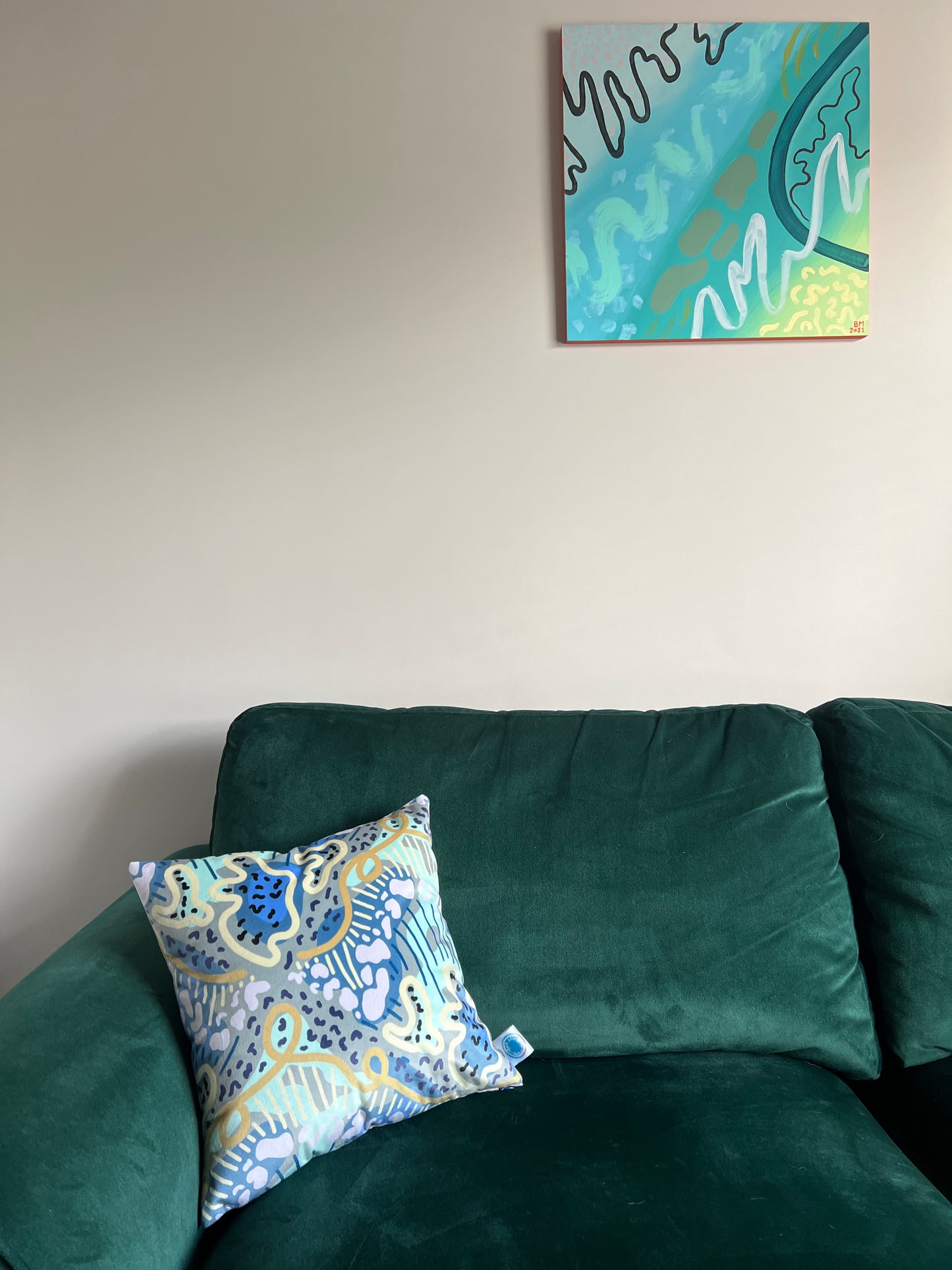 A statement cushion made up of organic shapes and lines in shades of greens, blues and yellows on a deep green sofa with an abstract painting on the wall above.