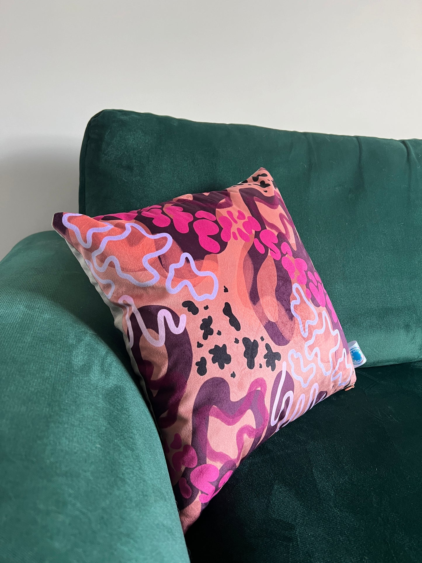 A colourful abstract cushion on a green velvet sofa. The cushion is made up of differing shades of orange, pink and purple in abstract shapes.