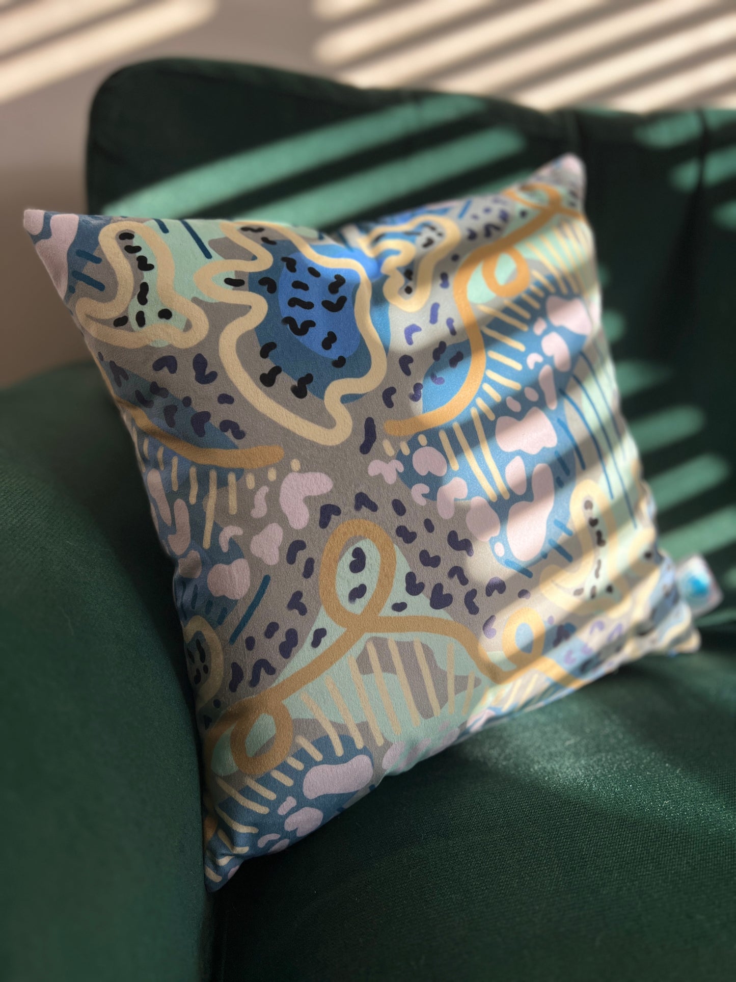 Blues, greens and yellows swirling lines and abstract shapes make up this cushion which is placed on a deep green sofa with stripes of sunlight illuminating part of the velvet fabric.